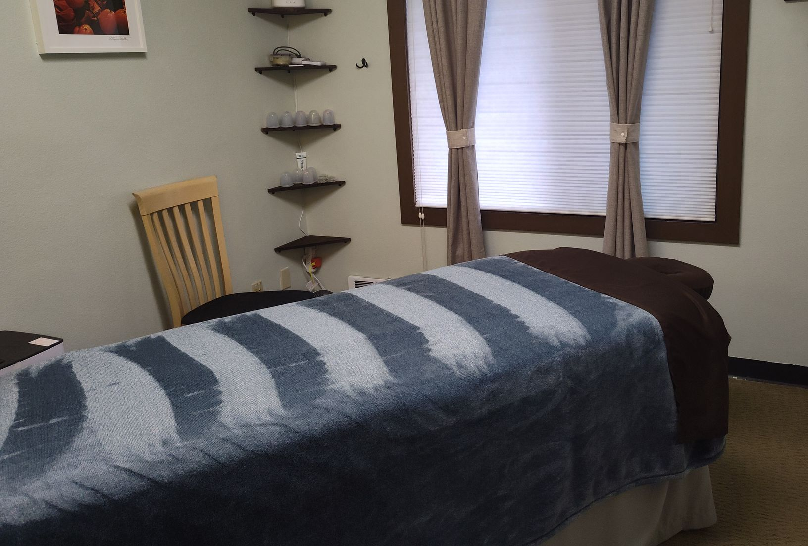 Massage room with bed, corner shelves, window with blinds, and chair.