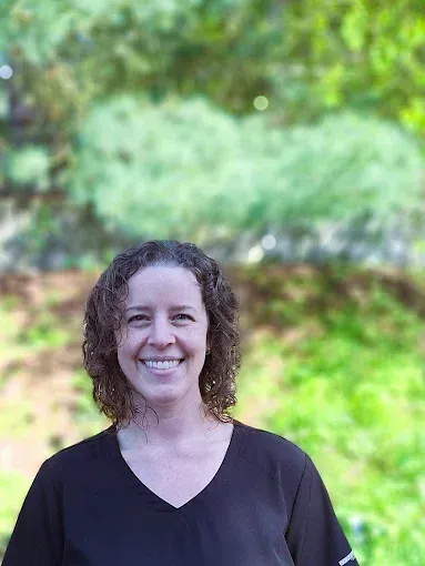 Woman with curly hair smiles in front of blurry green foliage.