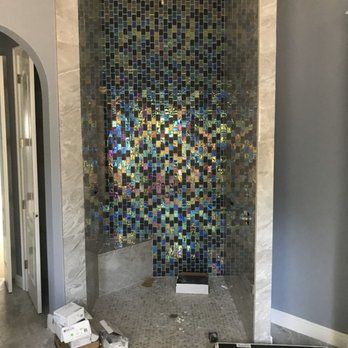 A shower niche with iridescent mosaic tile on the back wall and light gray tile on the floor and side walls. Boxes and debris are on the floor.