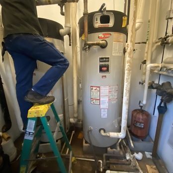 A person in blue jeans stands on a stepladder, working on a water heater. The setting is a utility room.