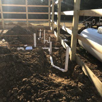 Plumbing pipes laid in a trench next to a wooden fence; a large white tank is visible in the background.
