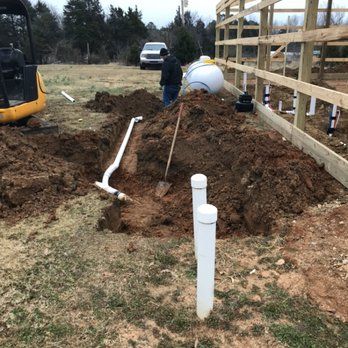 Construction site with exposed plumbing, dirt piles, and a man working. A small excavator and a white tank are also present.