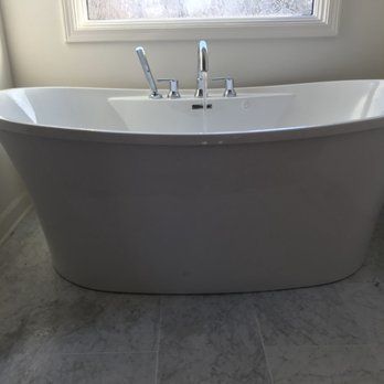 Freestanding white bathtub with chrome fixtures against a window; set on gray tiled floor.