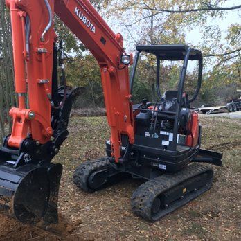 Orange Kubota mini excavator digging in a grassy area. The machine has black tracks, a cabin, and is set against a background of trees.