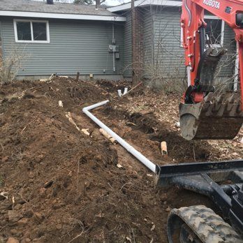 An excavator digs a trench in a yard next to a house, with white pipes visible in the ditch.