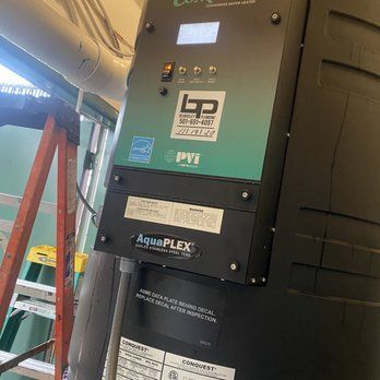 Black and green water heater control panel with digital display, on a large tank, next to a ladder.
