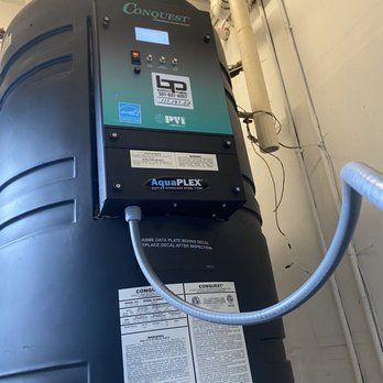 A black Conquest pool heater with control panel. A grey hose is connected to the side. The unit is in an enclosed space.