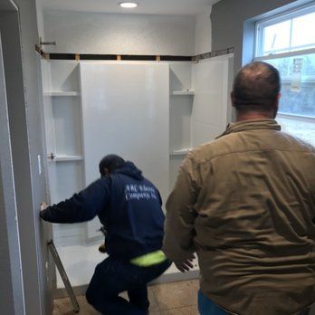 Two men installing a white shower stall. One man is crouching, working on the stall; the other stands nearby in a tan shirt. Bright, well-lit bathroom.