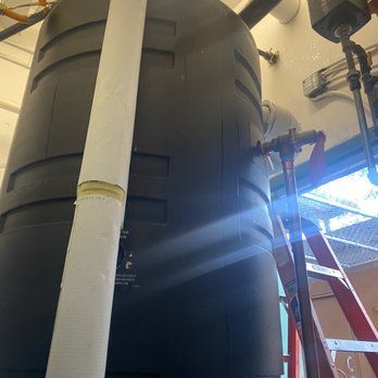 Large black water tank with attached pipes, a ladder, and a white pipe alongside. Indoors, possibly a utility room.
