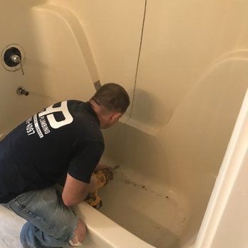 Man drills into a beige bathtub. He wears a dark shirt and jeans, focused on his work. The setting appears to be a bathroom.