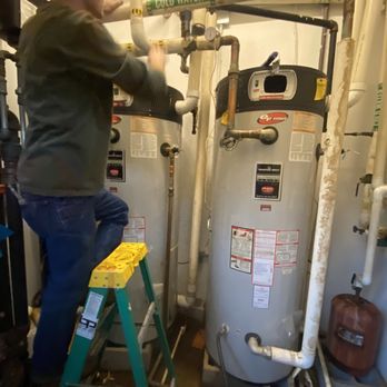 A person repairs plumbing near two large water heaters. They stand on a yellow step ladder in a utility room.