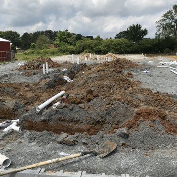 Construction site with exposed white plumbing pipes in trenches surrounded by dirt, shovel in foreground, under cloudy sky.
