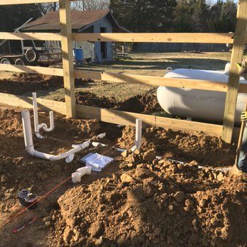 Construction site with exposed plumbing in a trench, next to a wooden fence and propane tank. Dirt and building materials are visible.