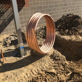 A coil of copper tubing sits in a dirt trench, near a brick wall and a support post, ready for installation.