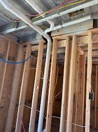 Interior view of a wood-framed wall with pipes and wires running along the ceiling and within the wall studs, unfinished construction.