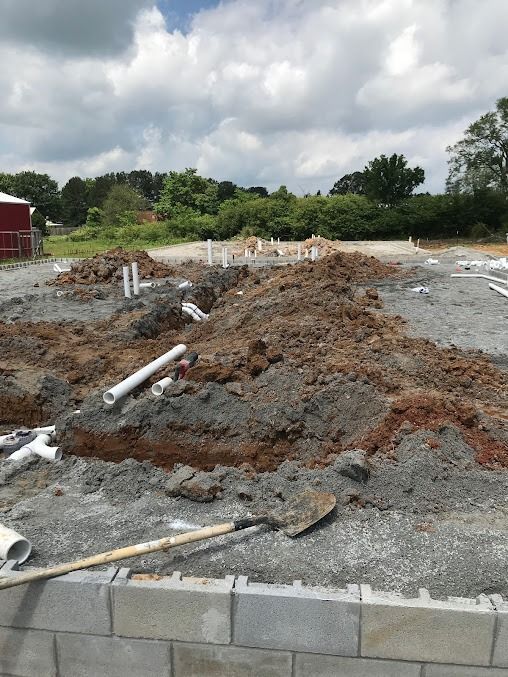 Construction site with exposed plumbing pipes and a partially built cinder block foundation; dirt and gravel surround the work.