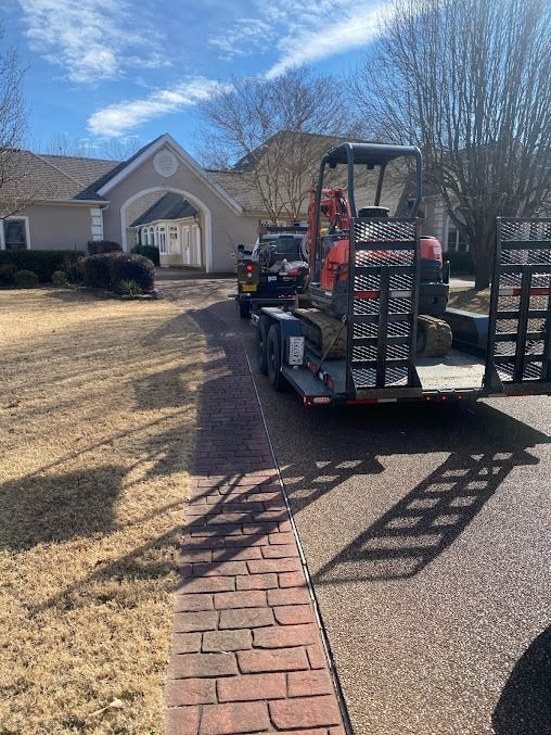 A small excavator on a trailer parked in front of a light-colored house on a sunny day.