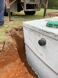 A concrete septic tank being installed in a trench. The tank is gray, with a green lid, near a construction site.