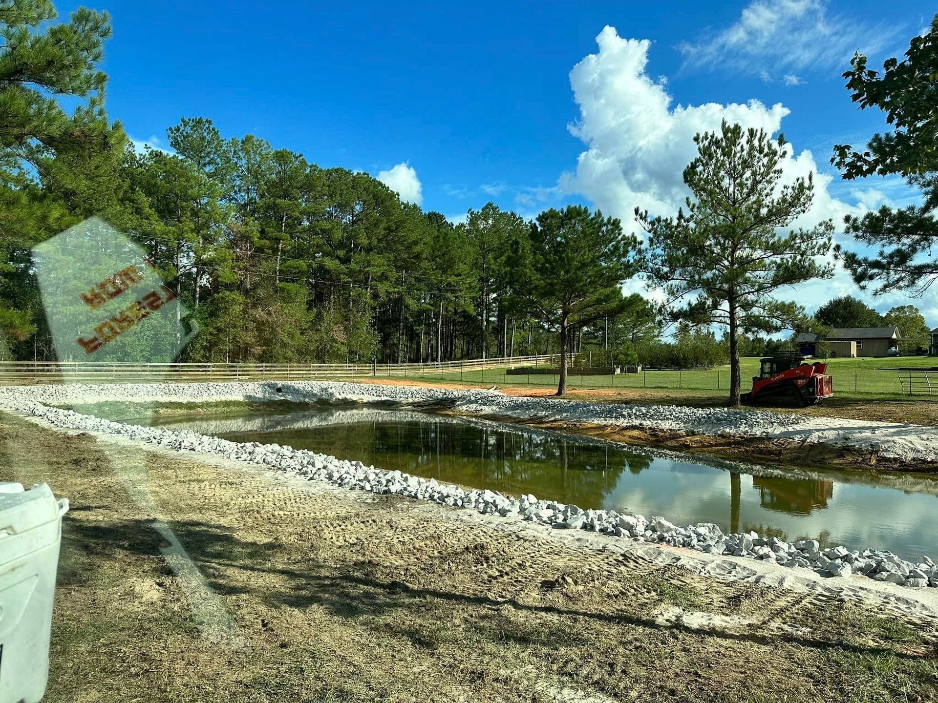 A newly constructed pond with a rock border is in front of a grassy area and trees under a blue sky. A tractor is visible.