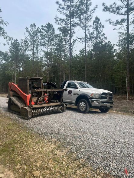 A red and black forestry mulcher next to a white truck on a gravel lot in a wooded area.
