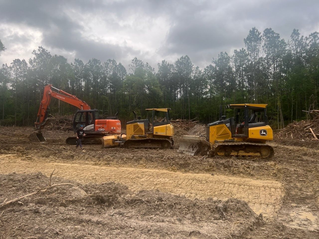 Three pieces of construction equipment: an excavator and two bulldozers on muddy ground, clearing a wooded area under a cloudy sky.