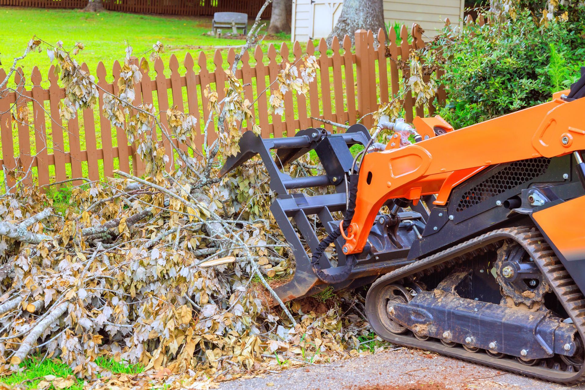 An orange skid steer loader with a grapple bucket clearing brush next to a wooden fence.