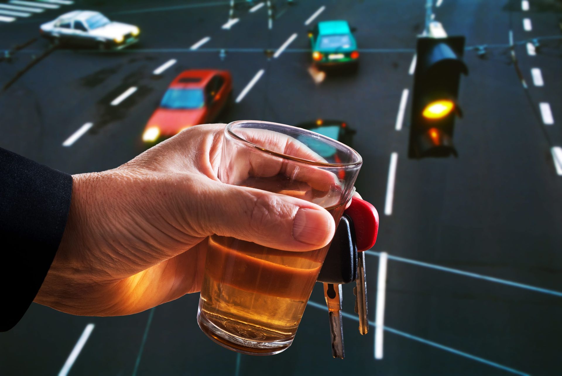 A person holds a glass of amber liquid and a set of car keys against a blurred background of a street intersection.