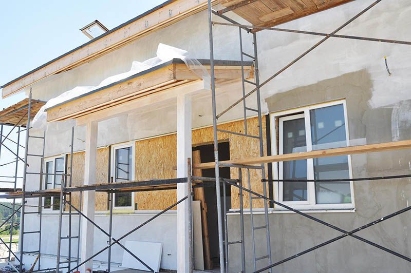 Extérieur du bâtiment en construction avec échafaudages. Stuc gris, fenêtres et boiseries visibles.