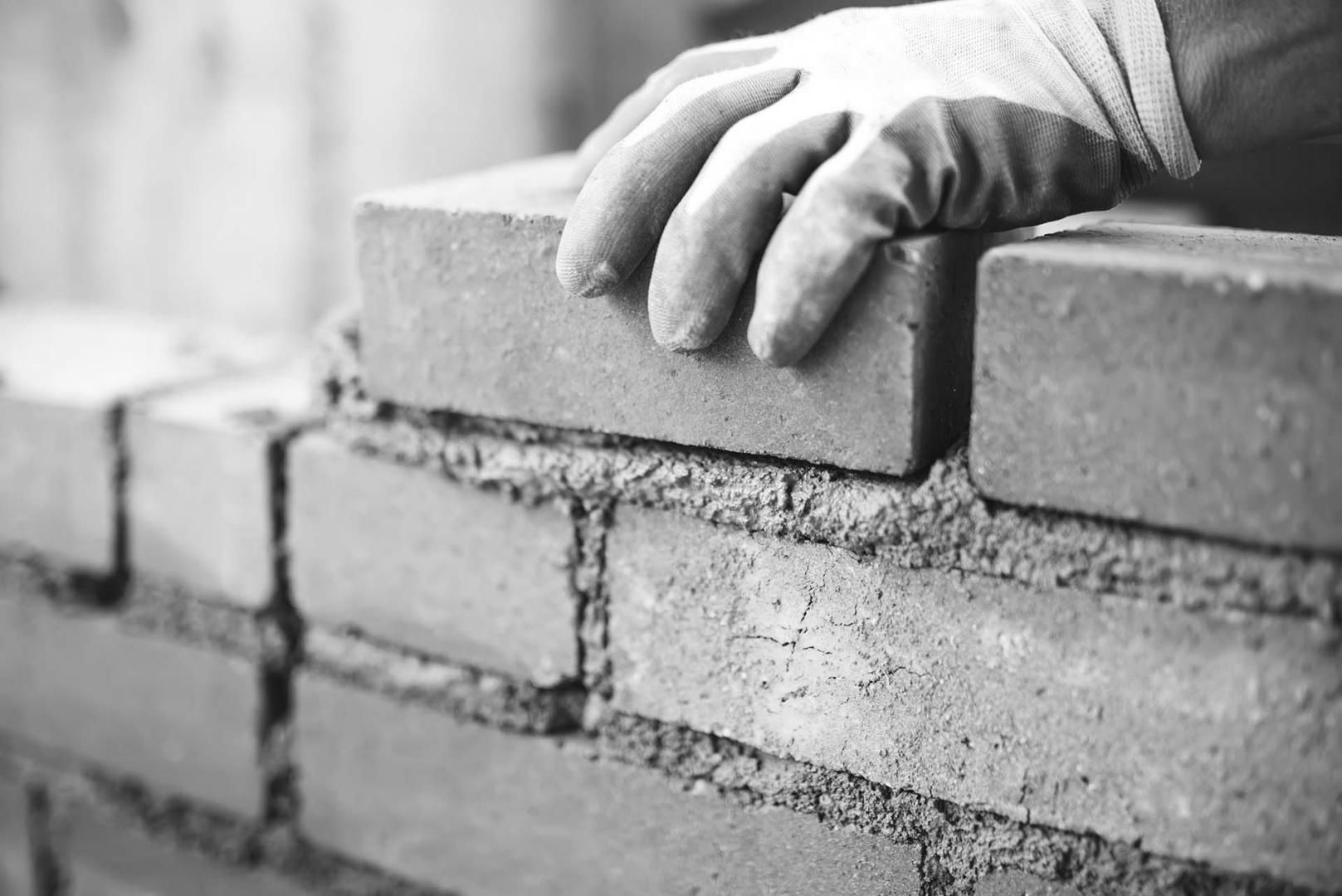 Main gantée posant une brique sur un mur de briques partiellement construit. Gros plan, noir et blanc.
