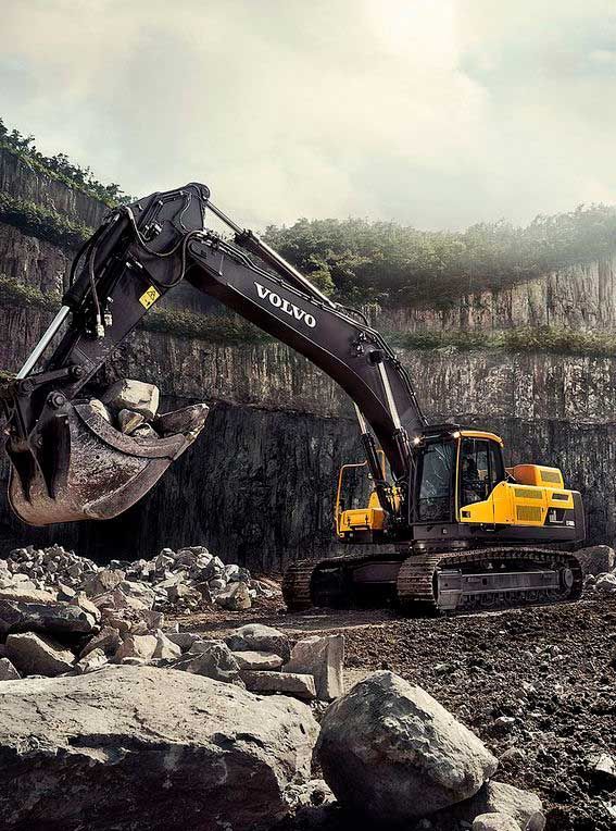 Pelle Volvo creusant des roches dans une carrière. Cabine jaune, bras gris foncé, rochers gris et falaise.