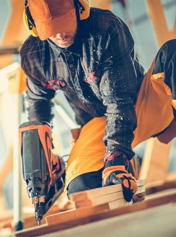 Ouvrier du bâtiment utilisant un pistolet à clous sur des planches de bois, portant un équipement de sécurité.