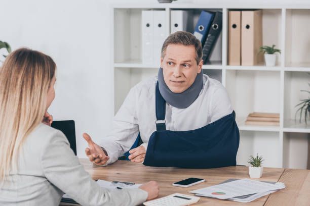 Man with neck brace and arm in a cast sits on a table in front of a blonde woman in an office.