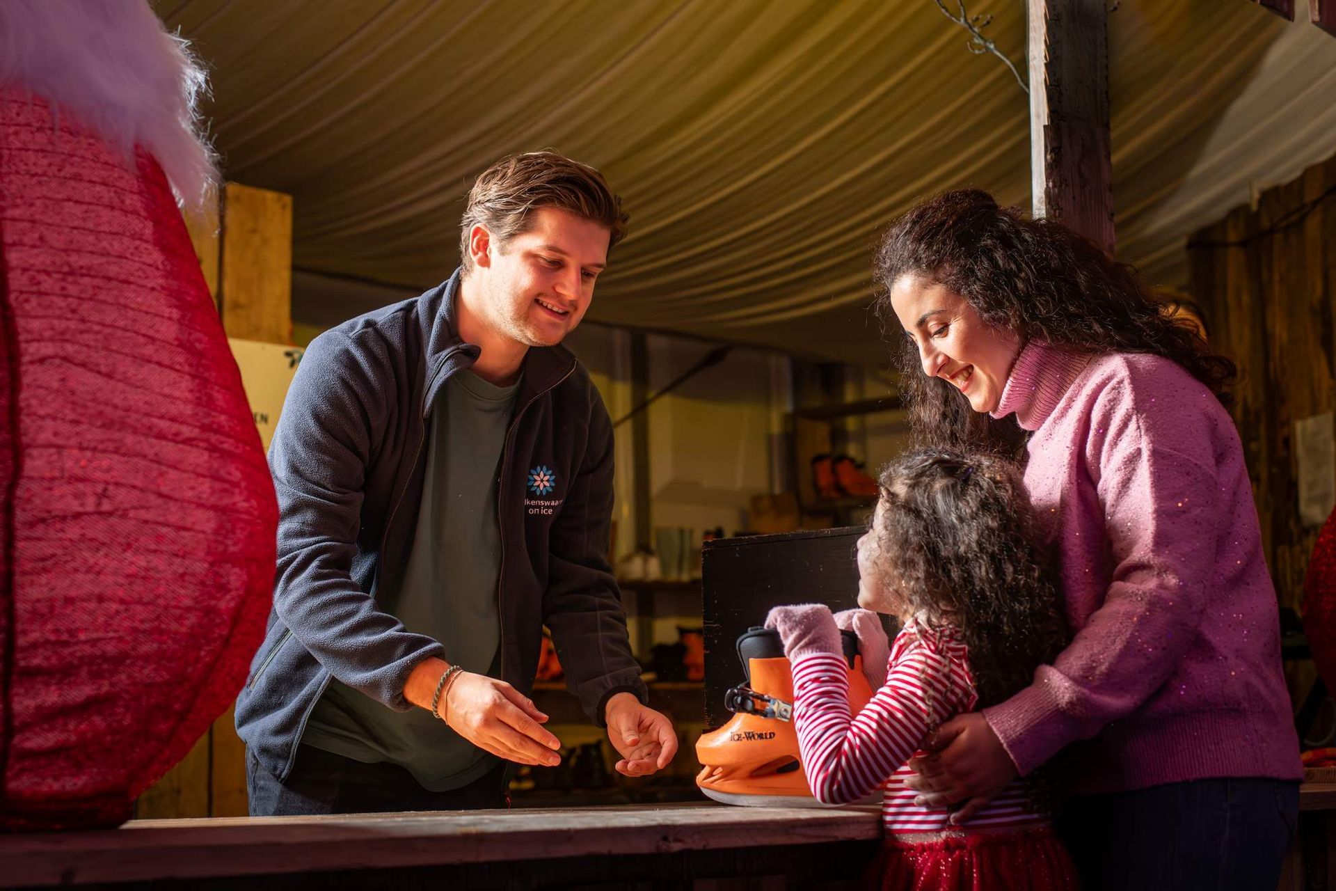 Een man helpt een kind en een vrouw bij een knutseltafel, met een oranje lantaarn in beeld. 