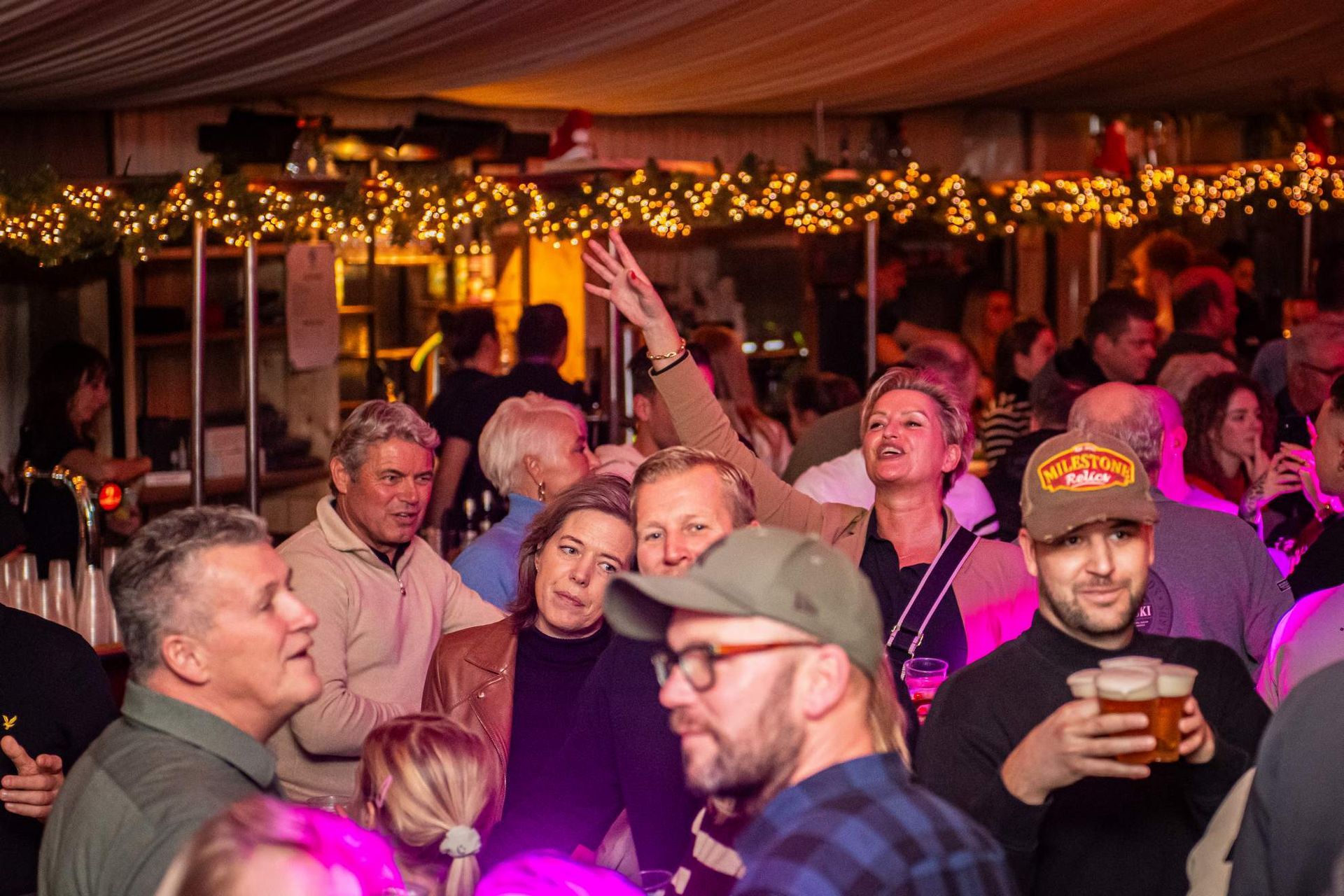 Mensen op een druk openluchtfeest, sommigen met een drankje in de hand, met lichtslingers boven hun hoofd.