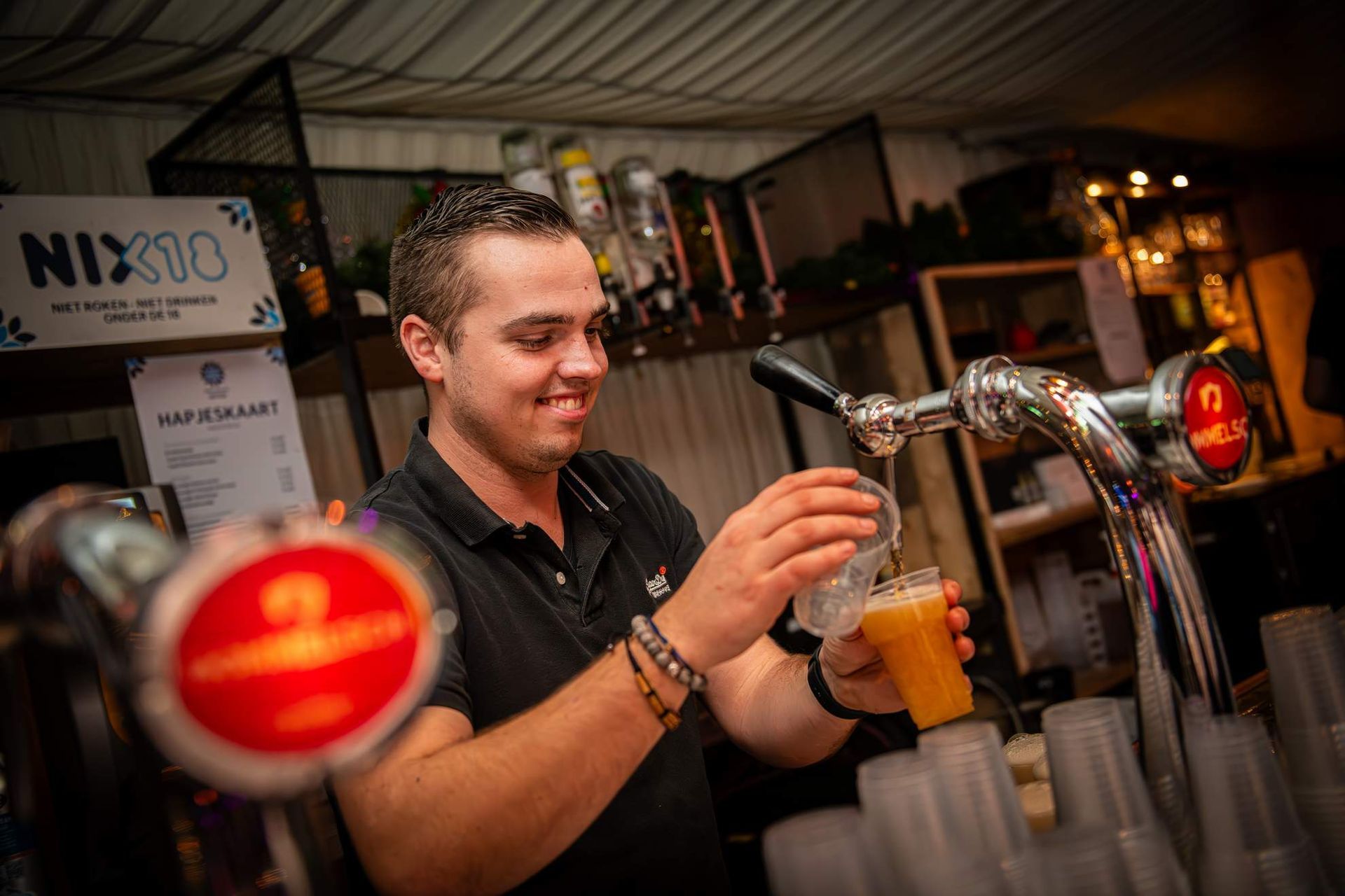 Barman schenkt bier van de tap in een plastic beker aan de bar; glimlacht.