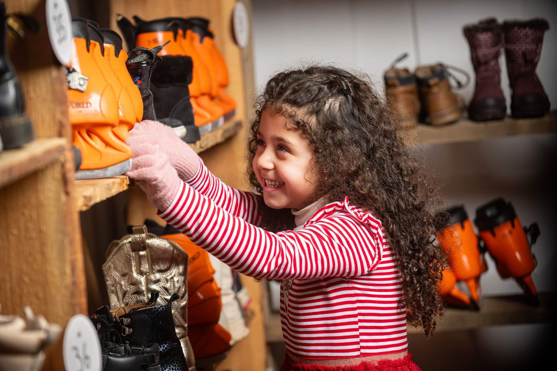 Een lachend meisje pakt oranje sneeuwlaarzen uit een houten plank in een winkel.