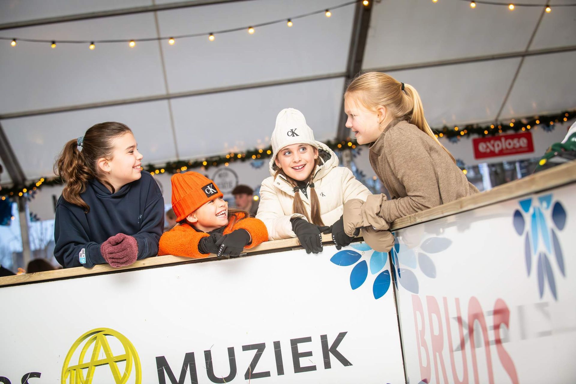 Kinderen leunen tegen een hek en kijken naar iets. Ze lachen en dragen een wintermuts en handschoenen.