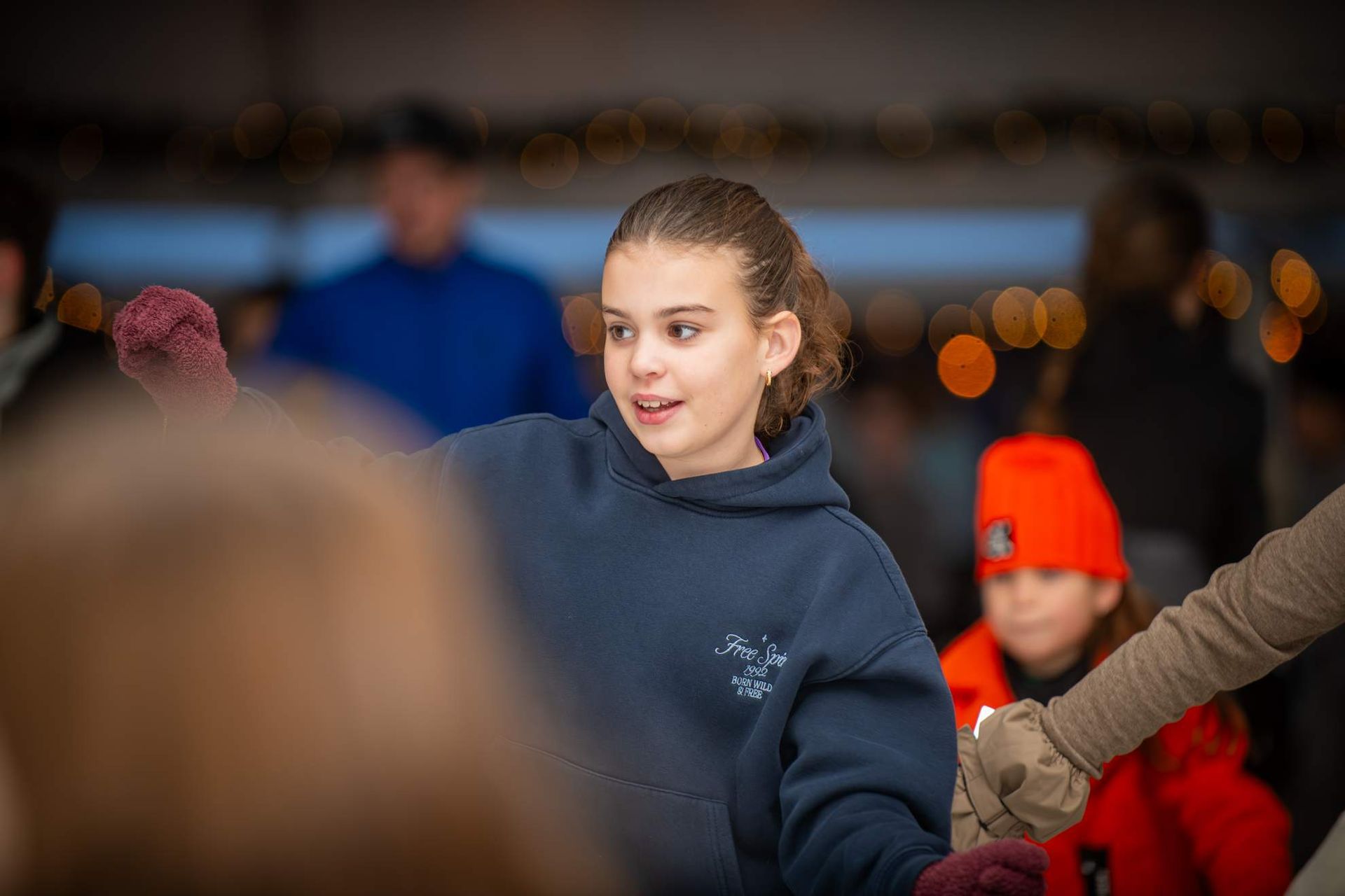 Meisje op schaatsen, gekleed in een blauwe hoodie en kastanjebruine handschoenen.