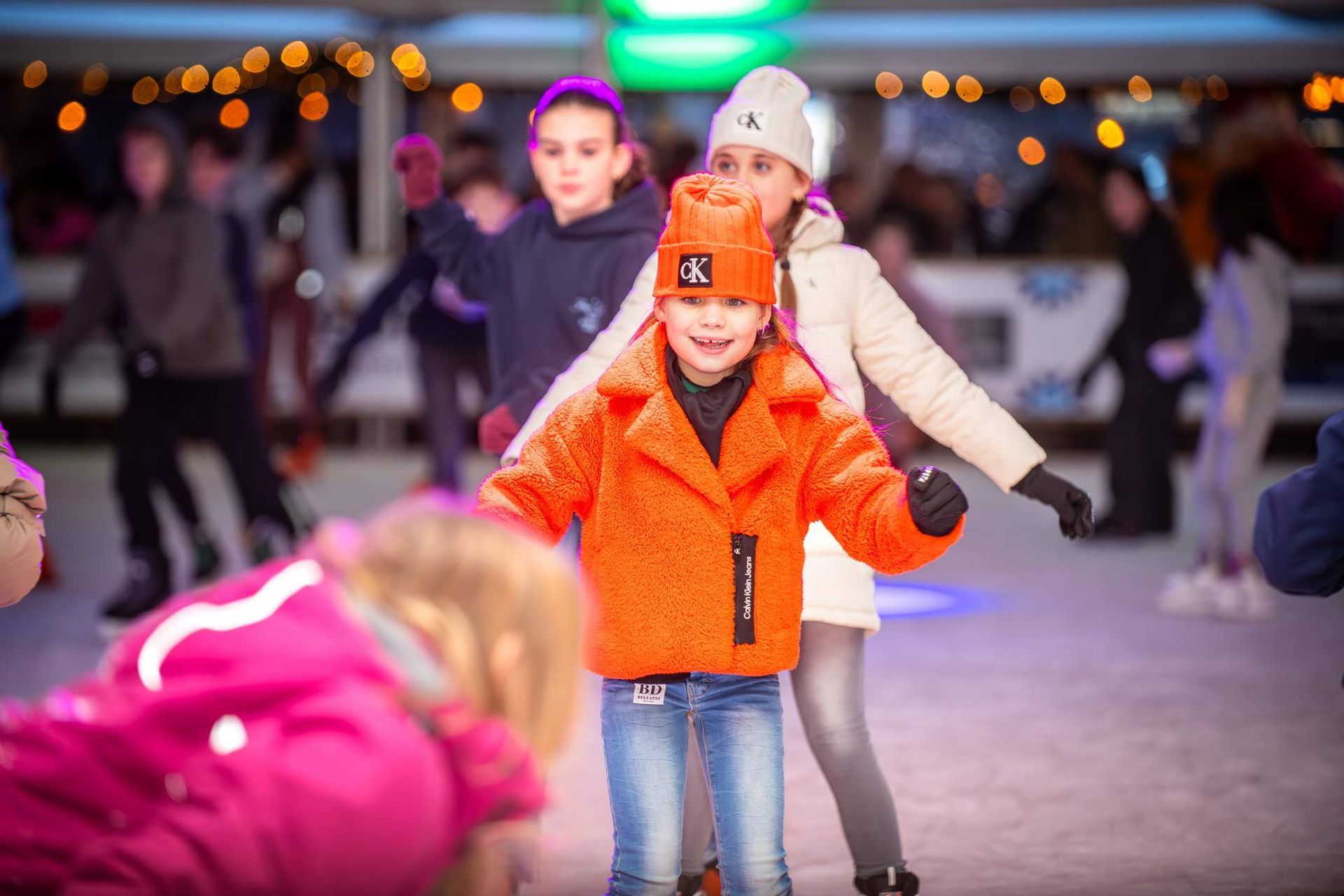 Kinderen zijn aan het schaatsen, één draagt ​​een feloranje jas en hoed, en lacht naar de camera.