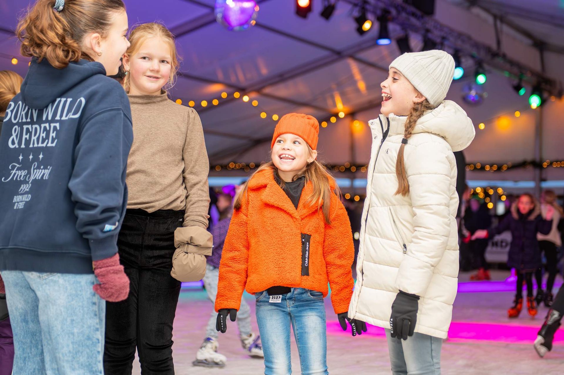 Vier lachende kinderen op een ijsbaan, onder een tent met lichtjes.