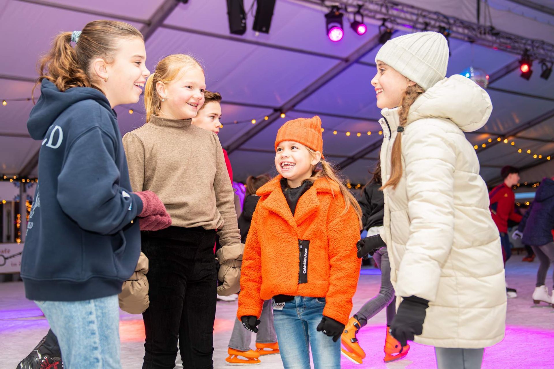 Een groep kinderen op schaatsen, pratend en lachend op een openluchtijsbaan met verlichting.