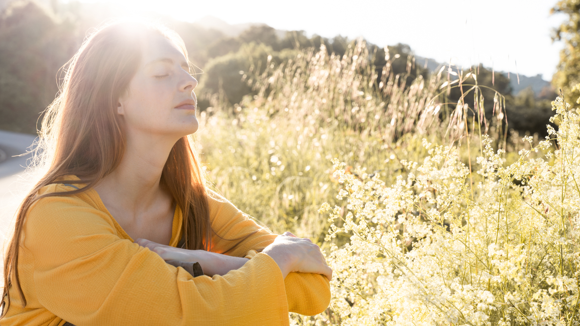 Woman in yellow shirt with eyes closed, sunlight filtering through tall grass.
