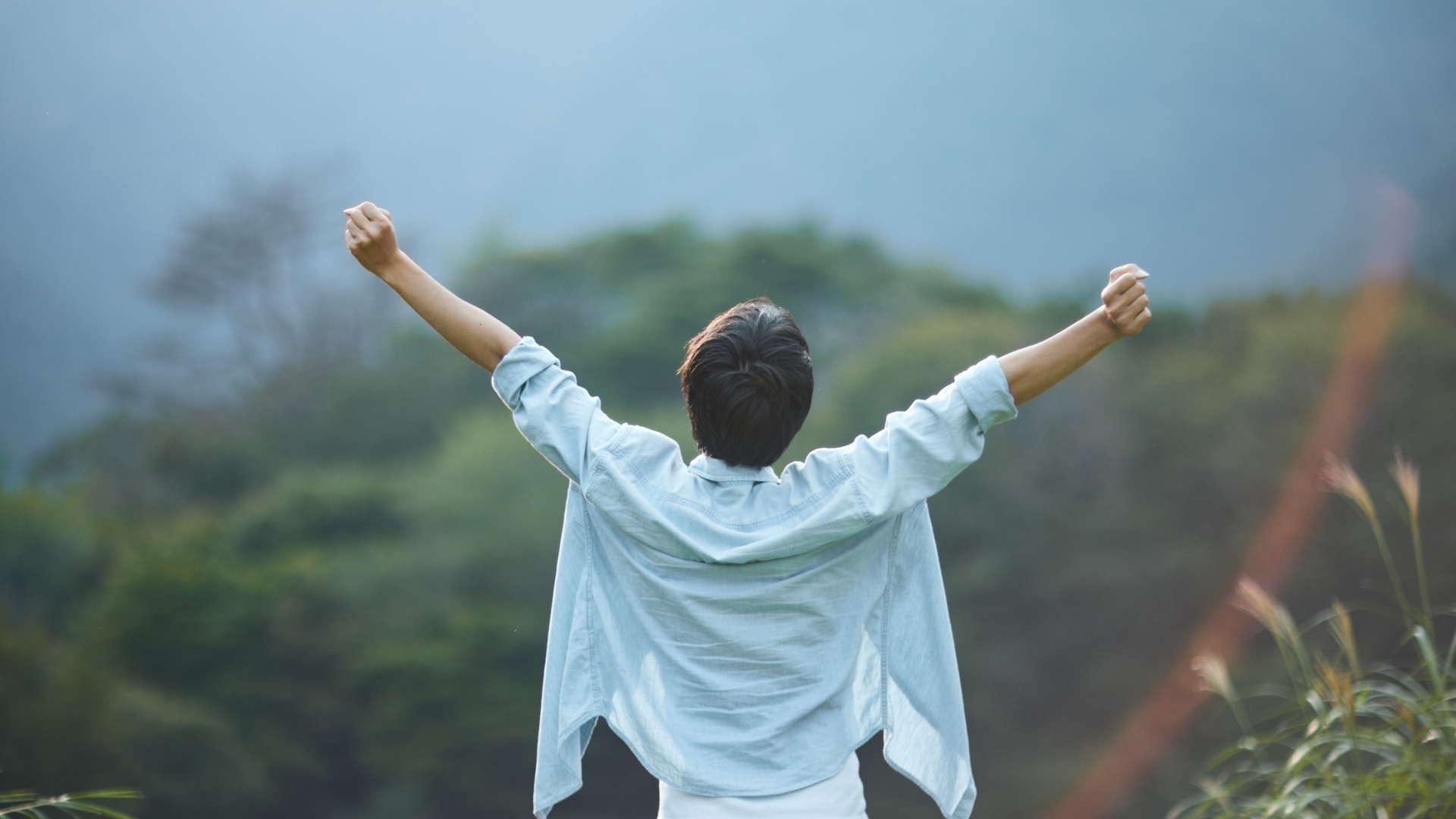 Person with arms raised in front of a blurred green landscape under a pale blue sky.