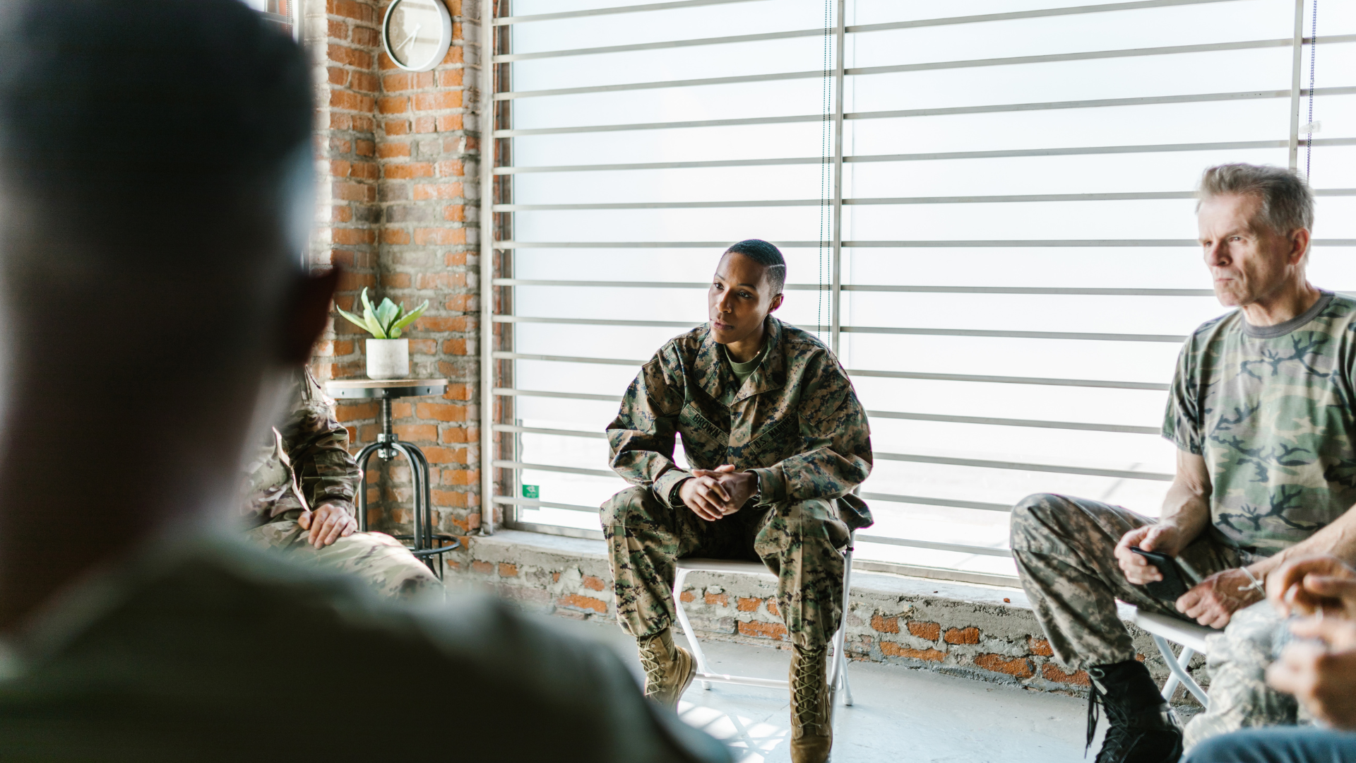 Soldiers in camouflage uniforms sit in a circle in a room with a brick wall.