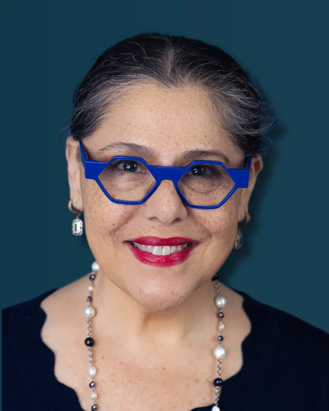 Woman with blue glasses, necklace, and red lipstick smiling, teal background.