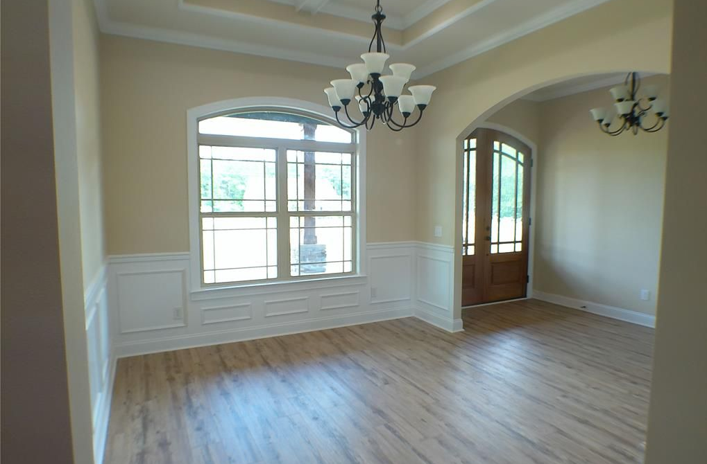Empty dining room with beige walls, white trim, wood floors, large window, and chandeliers.