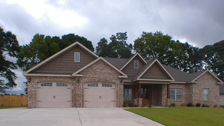 Brick house with two-car garage, brown roof, and green lawn under a cloudy sky.