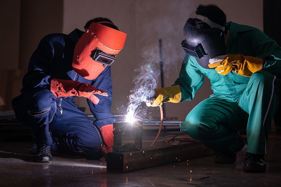 Two people in protective welding gear and uniforms working together to weld a piece of metal in a dim workshop.