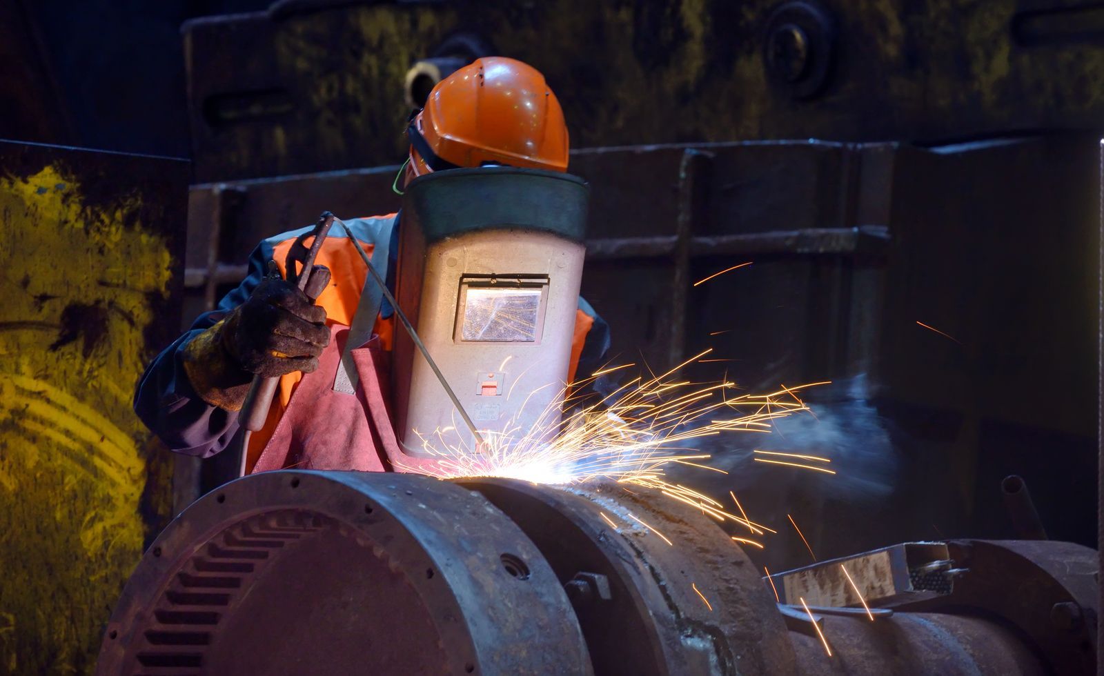 A worker in a hard hat and safety mask uses a welding torch on a large metal cylindrical part, creating sparks.