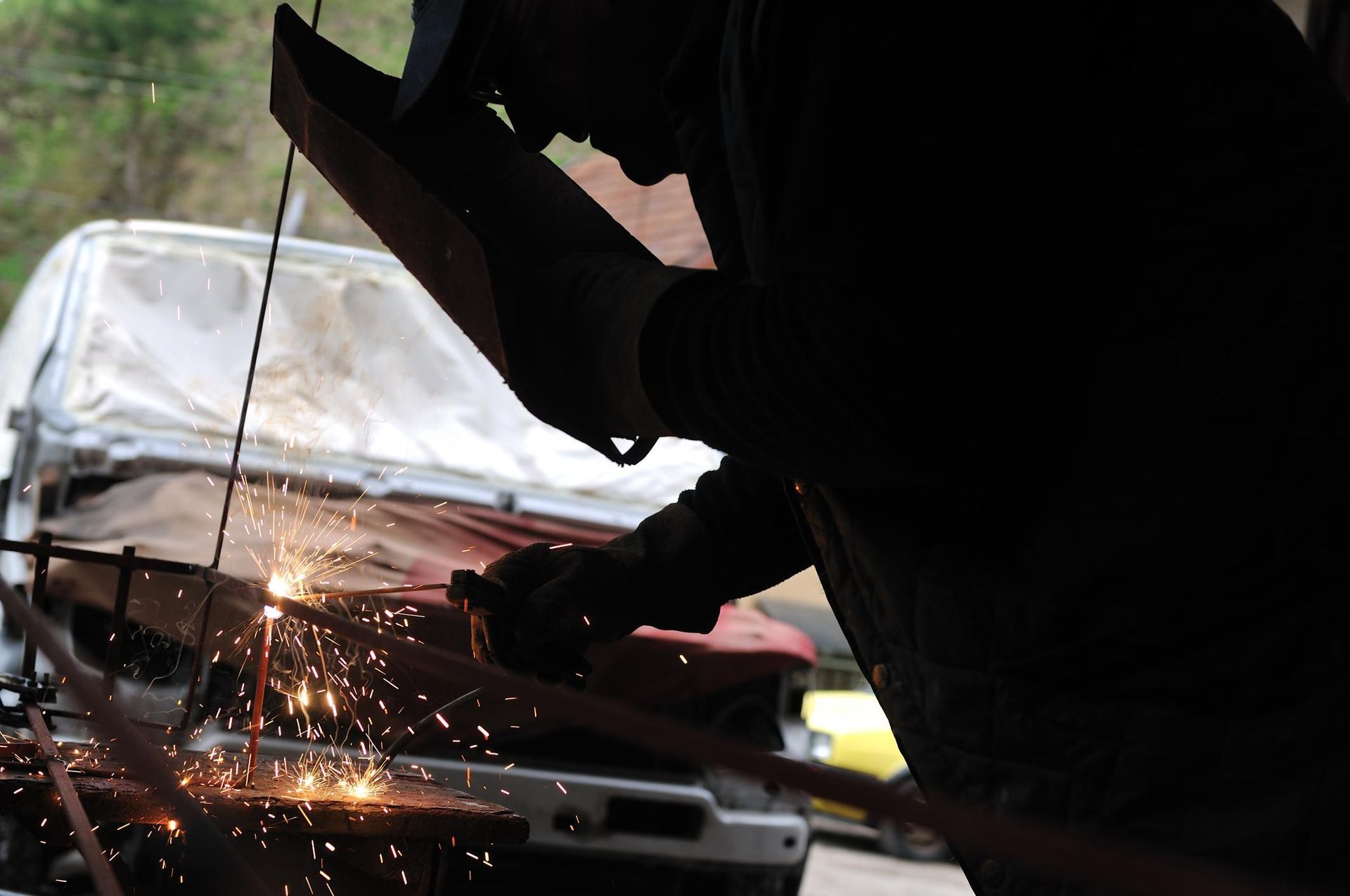 A welder wearing a protective mask works on metal, producing bright sparks in an indoor garage setting.